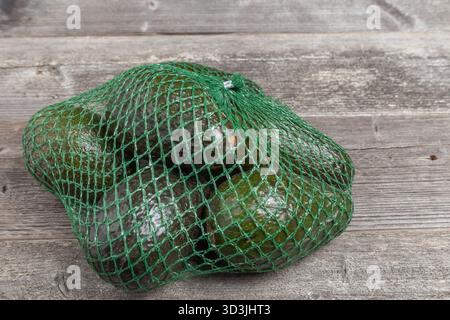 Close up of dark green avocados in a green mesh sack on weathered wood plank background. Stock Photo