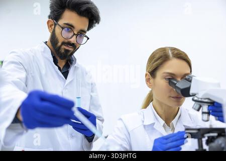 Young scientists using pipette, test tube and microscope in medical science laboratory, researching medical sample blue liquid together Stock Photo