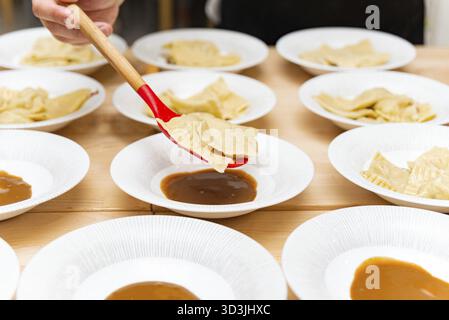 Food, A chef plates freshly cooked ravioli onto white dishes with sauce, using a red spoon, preparing an elegant pasta dish Stock Photo