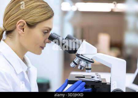 Side view portrait of blonde woman scientist looking biochemical cell in laboratory using microscope, investigating molecular biology Stock Photo
