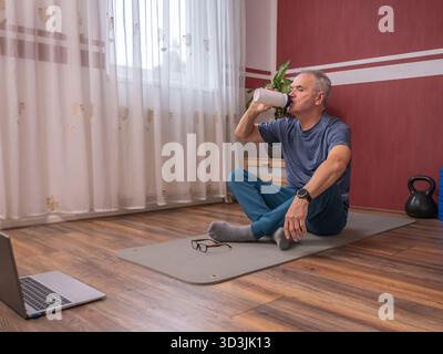 Senior man resting on yoga mat and drinking water after home workout Stock Photo