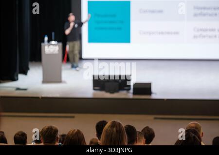 Speaker presents at conference stage with podium, screens display charts, audience seated listening attentively, microphones and water bottle on podium, professional event atmosphere Stock Photo
