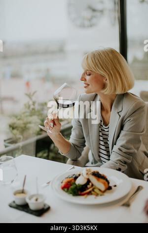 A woman in a gray blazer sips red wine while dining at an upscale restaurant. A plate of gourmet food is in front of her, and the city skyline is visi Stock Photo