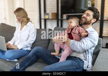 Father carrying sleeping daughter while looking at woman using laptop on sofa Stock Photo