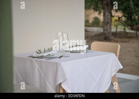 Elegant outdoor table set for two, featuring white plates, glassware, and a simple olive branch on each napkin. Stock Photo