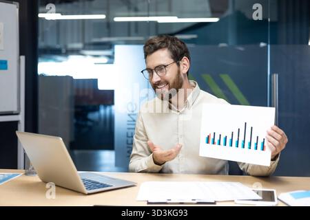 A young man, a company director and specialist, holds an online meeting with partners and clients, talks online and shows graphs and work dynamics to the camera. Stock Photo