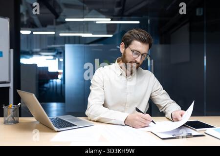 A serious and focused young male businessman is sitting in the office at his desk and working with documents. Stock Photo