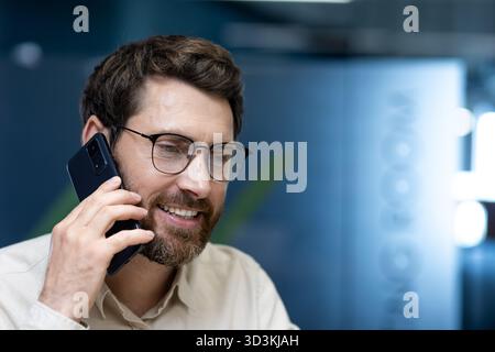 Close-up photo of a young man in glasses and with a beard talking on the phone at his workplace in the office. Stock Photo