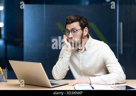Tired young male office worker sitting at his desk, leaning his head on his hand and looking boredly at the laptop screen. Stock Photo