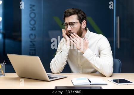 A tired and overworked young man sits in the office at a desk with a laptop and yawns. Stock Photo