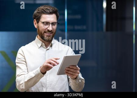 Smiling fashionable male programmer, office worker standing indoors and using a tablet in his hands. Stock Photo