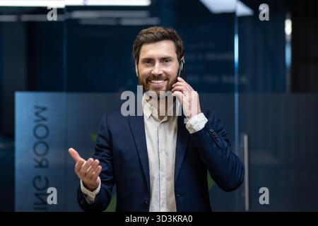 Close-up portrait of a young businessman talking on the phone in the office, gesturing with his hands and looking at the camera. Stock Photo