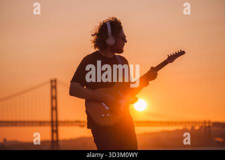Hipster street musician in black playing electric guitar in street outdoors on sunset with lens flare and light leak Stock Photo