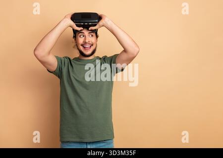Young man wearing a green t shirt smiles while wearing a virtual reality headset against a beige background in a casual lifestyle advertising scene Stock Photo