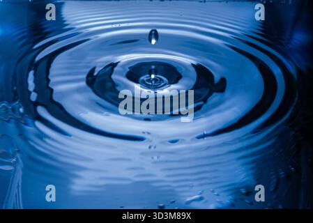 226.Droplet falls into water forming splash and concentric ripples. Deep blue reflections highlight fluid motion, surface tension, and wave propagatio Stock Photo