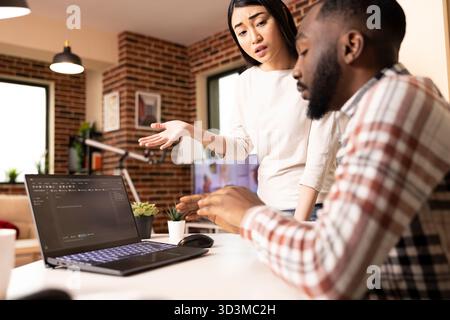 Diverse couple collaborating on laptop project in modern brick wall home. Asian girlfriend gestures while african american boyfriend listens, highlighting positive teamwork in warm light. Stock Photo