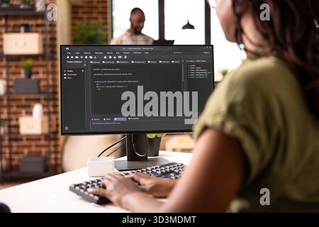 Young female software developer typing code on computer while working on startup project remotely. Black woman using desktop pc and programming new web application from modern apartment. Stock Photo