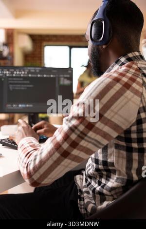 Male software developer with headphones coding from home, using computer to review programming scripts. African american it freelancer listens to music and works on tech solutions in modern apartment. Stock Photo