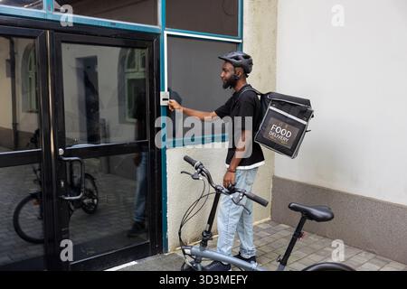 Food delivery worker with a backpack approaching a front door, delivering restaurant takeaway meal order to a client. Express courier delivery at the doorway, using door bell intercom. Stock Photo