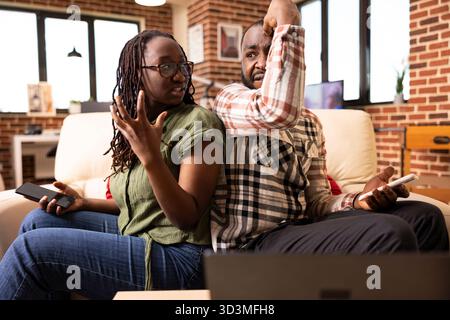 Frustrated black couple, each holding cellphone and arguing with expressive hand gestures. Man and woman sit back to back on couch having relationship problems and communication issues at home. Stock Photo