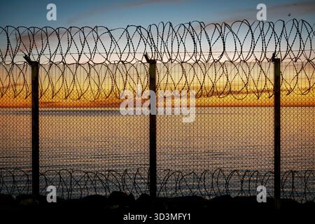 A captivating silhouette of razor wire and a chain-link fence against a vibrant sunset over the sea. Concertina wire. Security, barrier, border, freed Stock Photo