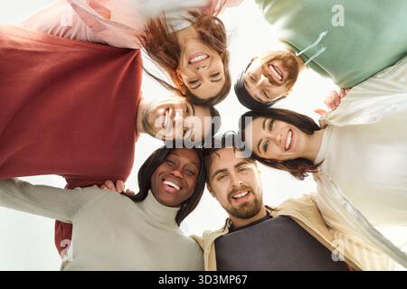 Smiling diverse group of friends standing in circle, looking down at camera and hugging each other. Stock Photo