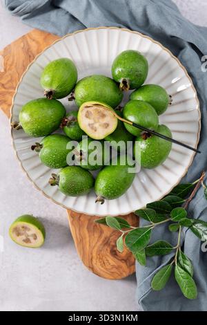 A close-up, top-down shot of a group of fresh, green feijoa fruits arranged on a white fluted plate. One of the fruits is cut in half, revealing its p Stock Photo