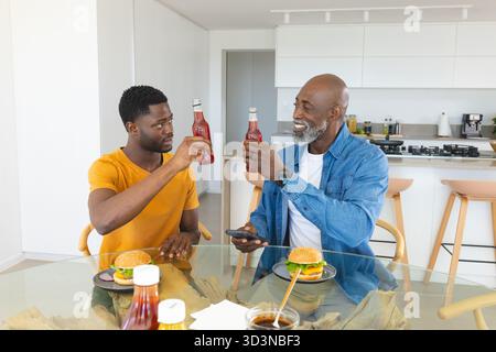 African American father and son celebrating lunch in kitchen, holding red bottles and hamburgers Stock Photo
