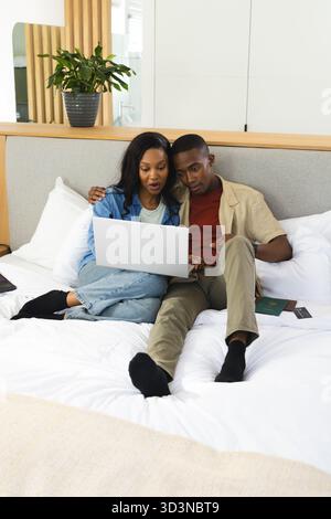 Lounging African American couple sharing laptop on bed in bedroom, with passport smartphone Stock Photo