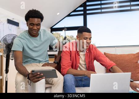 Collaborating Diverse male friends working on couch in modern home office, using tablet and laptop Stock Photo
