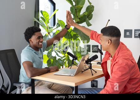 High-fiving Diverse male colleagues sharing podcast setup at office, with microphones and laptop Stock Photo
