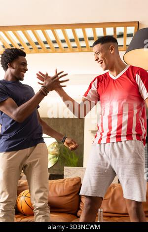 High-fiving Diverse male friends on leather couch in modern living room, with basketball Stock Photo