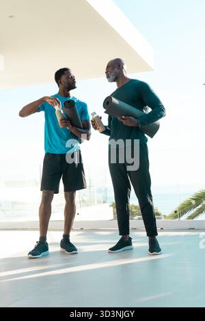 Standing African American family holding yoga mats and water bottles on terrace overlooking ocean Stock Photo