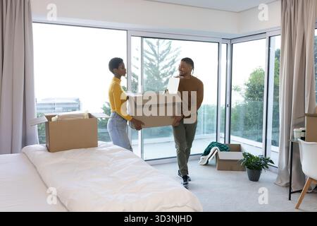 Diverse couple carrying large cardboard box in modern bedroom, with potted plant and white bedding Stock Photo