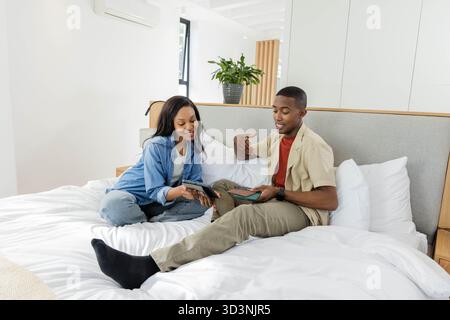 African American couple using digital tablets sitting on bed in bedroom, with smartphone and plant Stock Photo