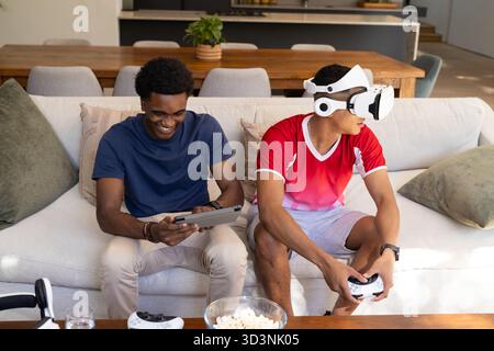 Diverse male friends playing games on sofa in living room, with tablet, VR headset and controllers Stock Photo