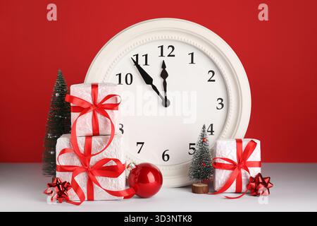 White clock with Christmas decorations and gift boxes on grey table against red background Stock Photo