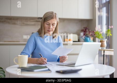 Mature woman reviewing financial documents and making notes while using a laptop and calculator for budget planning and remote work from her modern kitchen Stock Photo