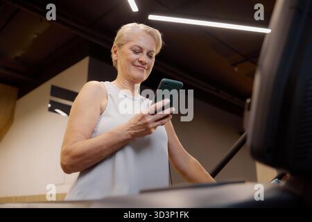 Caucasian Senior Woman Using Smartphone While Exercising on Treadmill Indoors Stock Photo