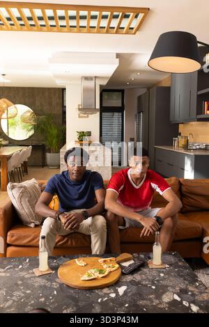 Sitting diverse male friends on brown leather couch in open-plan living room, with pizza board Stock Photo
