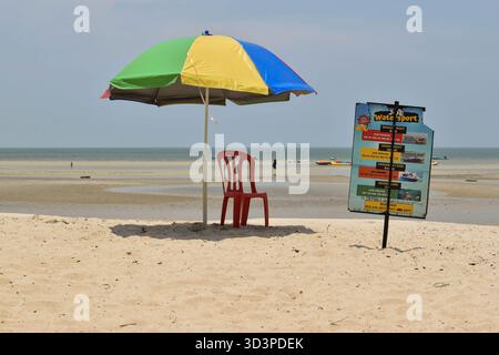 Multicolored umbrella shades red plastic chair on quiet beach; nearby signboard advertises water sports, highlighting tourism and coastal recreation. Stock Photo