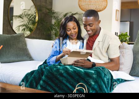 Smiling African American couple examining gift box on sofa in living room, holding notecard and pen Stock Photo