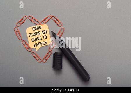 Black pen rests beside a heart made of paperclips, featuring a motivational note that reads good things are going to happen, set against a smooth gray Stock Photo