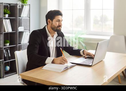 Focused businessman working on laptop and taking notes at desk while planning tasks in office. Stock Photo