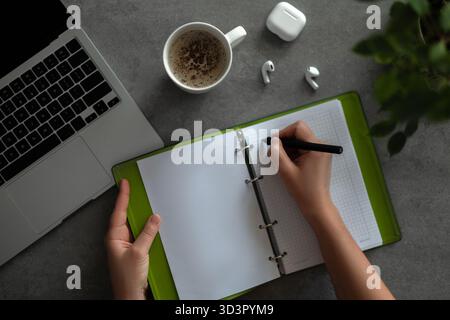 Top view of a cropped unrecognizable woman writing in a notebook, with a laptop, coffee cup, and AirPods nearby, embodying a productive home office se Stock Photo