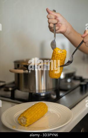 Cropped unrecognizable preteen girl at home in the kitchen cooking corn in a saucepan. She uses utensils to serve boiled corn on the cob onto a plate, Stock Photo