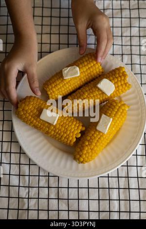 Top view of a cropped unrecognizable preteen girl at home in the kitchen, arranging corn cobs with butter on a plate, highlighting culinary skills and Stock Photo