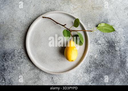 Top view of a yellow pear with green leaves on a textured gray plate set against a concrete background, exuding minimalism and natural elegance. Stock Photo