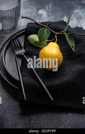 Yellow pear placed on a black cloth napkin with green leaves, set on a dark ceramic plate. Black fork and a glass are beside it, creating a minimalist Stock Photo