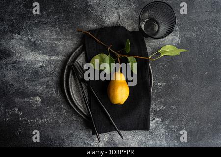Top view of a stylish table setting featuring a single yellow pear on a black napkin, gray ceramic plate, and matte black utensils, all on a textured Stock Photo